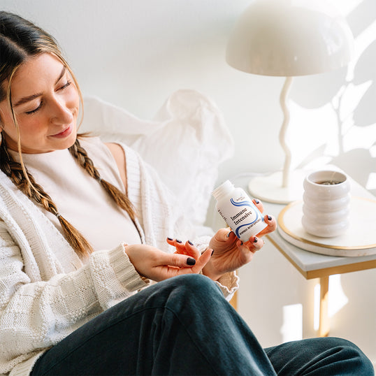 A woman in a cozy indoor setting holds a white bottle of 'immune support' capsules. She is wearing a white knit cardigan and dark pants, sitting near a white table lamp and a white ceramic cup on a side table.