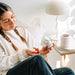 A woman in a cozy indoor setting holds a white bottle of 'immune support' capsules. She is wearing a white knit cardigan and dark pants, sitting near a white table lamp and a white ceramic cup on a side table.
