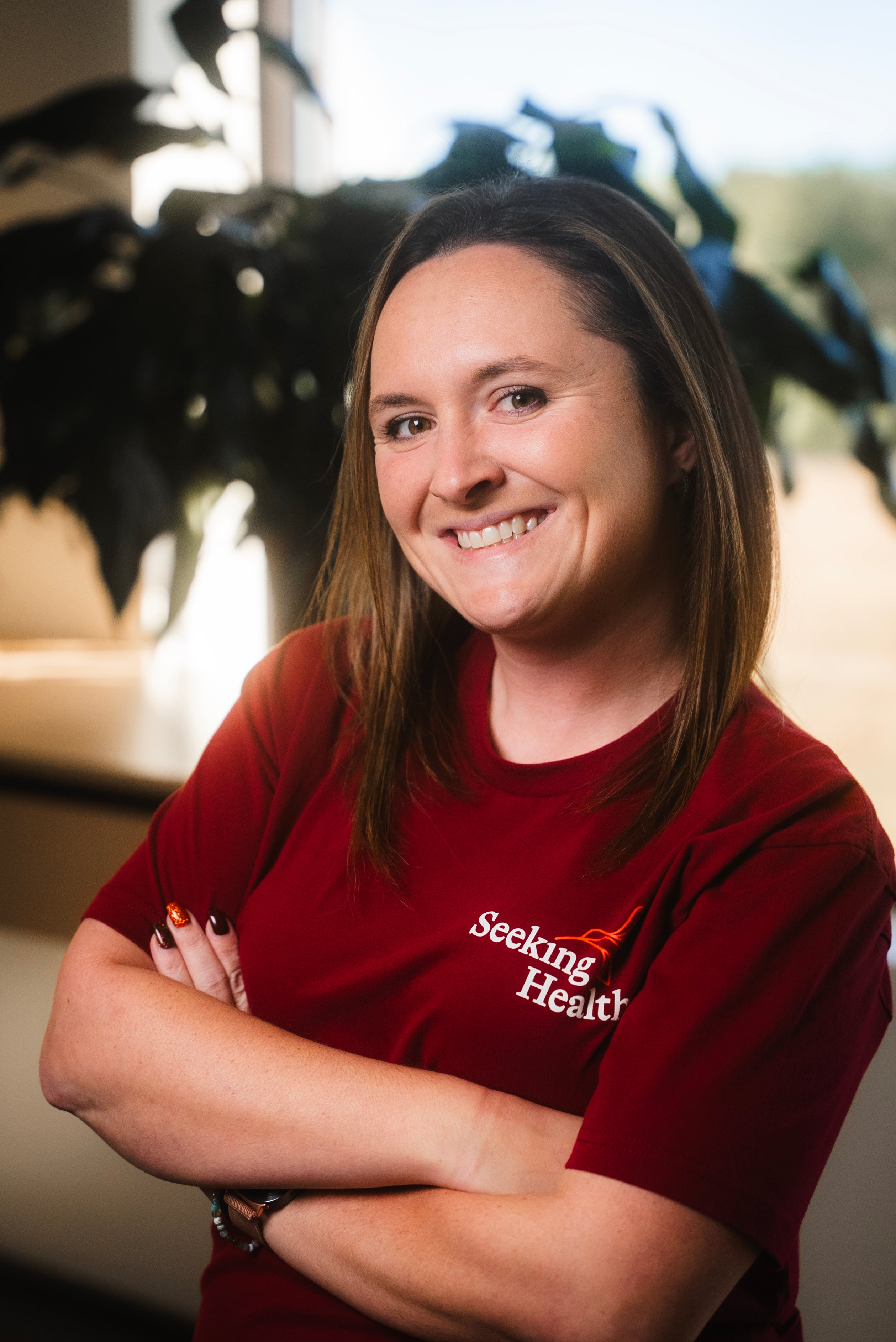 Person wearing a red 'Seeking Health' shirt with arms crossed, blurred plant and indoor setting in the background