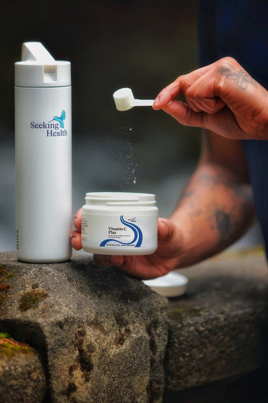 A person's tattooed hands are shown holding a white scoop over a jar of Seeking Health Vitamin B3 powder, with powder visibly falling from the scoop. A tall white bottle of Seeking Health is standing on a stone surface in the background.