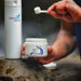 A person's tattooed hands are shown holding a white scoop over a jar of Seeking Health Vitamin B3 powder, with powder visibly falling from the scoop. A tall white bottle of Seeking Health is standing on a stone surface in the background.